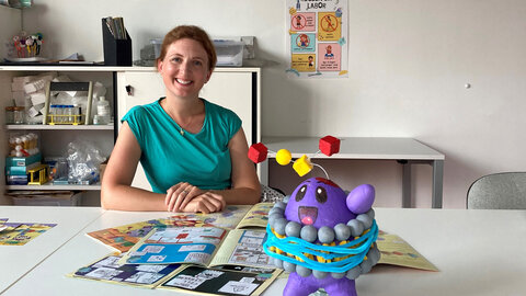 Young researcher sits at the desk on which comics lie and a homemade purple protein with red antennae for illustration for children