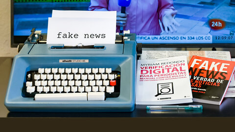 Fake News mit Schreibmaschine geschrieben, daneben liegen Sachbücher, im Hintergrund ein TV-Bildschirm 