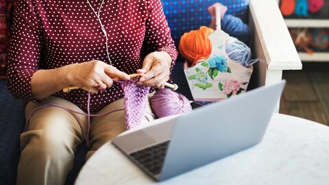 Upper body of an elderly woman knitting and sitting in front of a laptop